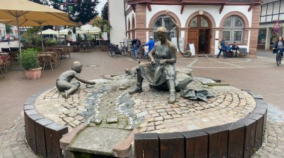 Der von Siegfried Speckhardt (geb. 1937) gestaltete Tabakbrunnen wurde 1991 zum Hessentag in Lorsch aufgestellt. Der begehbare Brunnen steht direkt vor dem alten Rathaus und zeigt eine Tabaknäherin bei der Arbeit. Der Brunnen erinnert an die mühevolle Arbeit der Forscher Tabaknäherinnen, die (bis zur Einführung der Tabaknähmaschinen ab 1956) als Vorbereitung für die Trocknung die Tabakblätter noch per Hand mit Nadel und Faden zu einem "Bandelier" zusammennähten und so bündelten.
Doch die Arbeit brachte auch etwas Gutes:
das Tabakläden war damals ein geselliges Ereignis. In der Runde der Näherinnen wurde viel erzählt, die letzten Neuigkeiten verbreitet und gesungen. Auch die Alten und Kinder halfen damals mit. Der B Brunnen erinnert also nicht nur an die aufwendige Arbeit, sondern auch an diesen schönen Brauch.